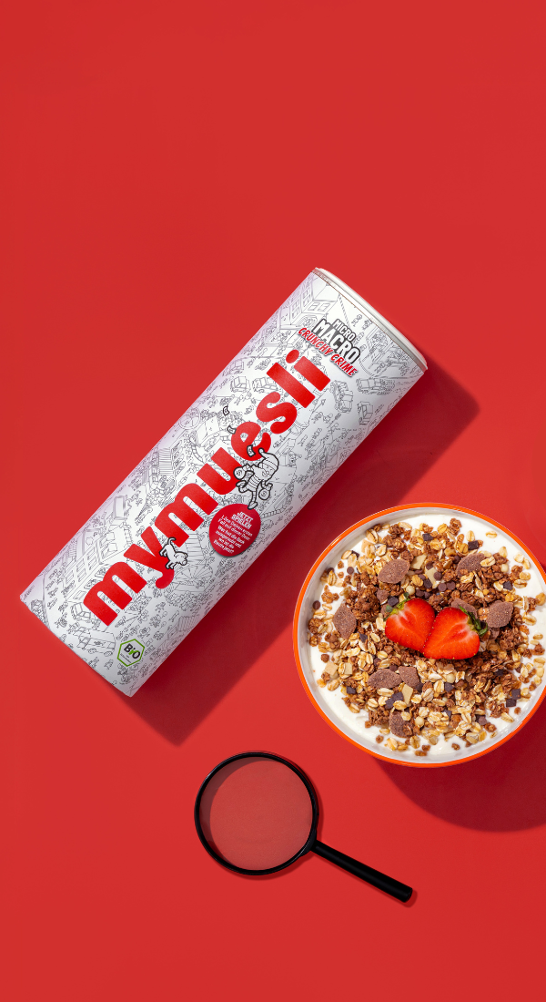 Das mymuesli Micro Macro Crunchy Crime Müsli mit integriertem Rätselspiel auf der Müslidose. Nebenan das angerichtete Super Choc Müsli mit frischen Äpfeln und Blaubeeren.