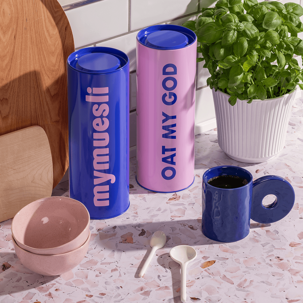 Zwei Vorratsdosen blau & rosa stehend neben einer Tasse und Schalen