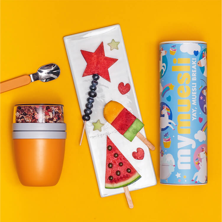 Fruchtspieße für Kinder auf einem Teller mit einem Löffel, 2go Becher und Müslidose daneben