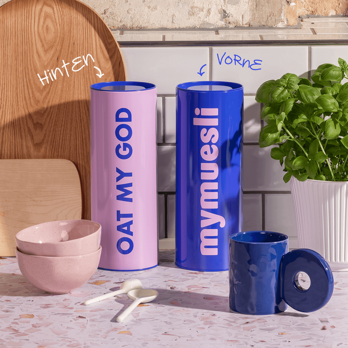 Zwei Vorratsdosen blau & rosa stehend neben einer Tasse und Schalen