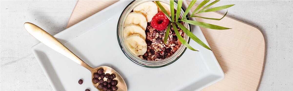 Banane Schoko Overnight Oats in einem Glas mit Bananenscheiben und einem Löffel daneben