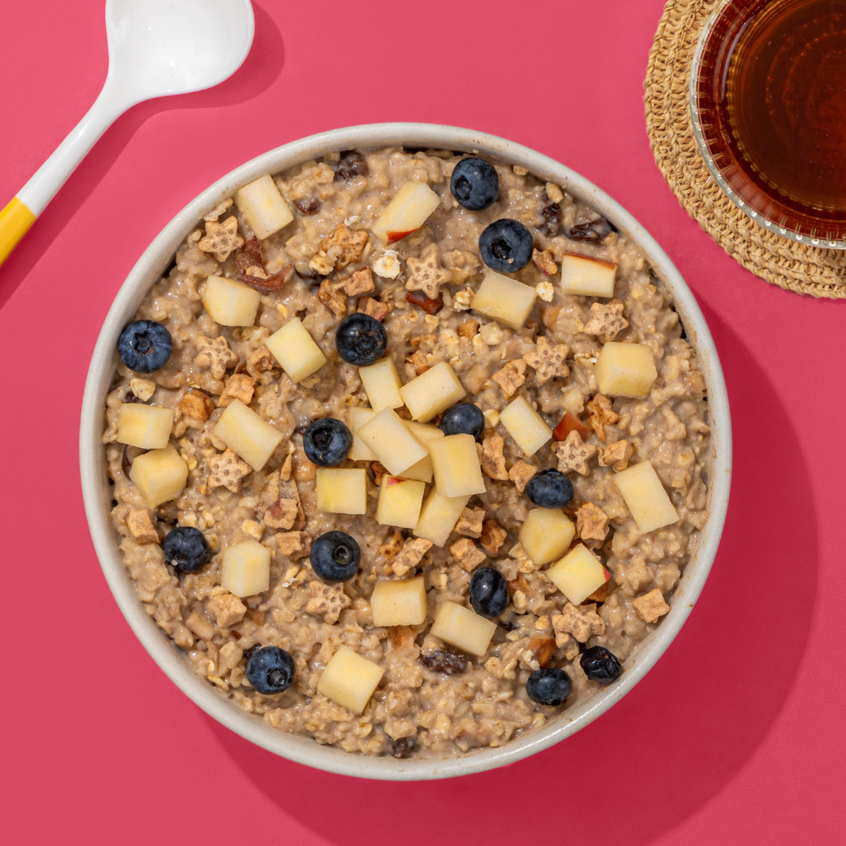 Porridge mit Blaubeeren und Apfelstücken in Müslischale, daneben ein Löffel und eine Tasse Tee