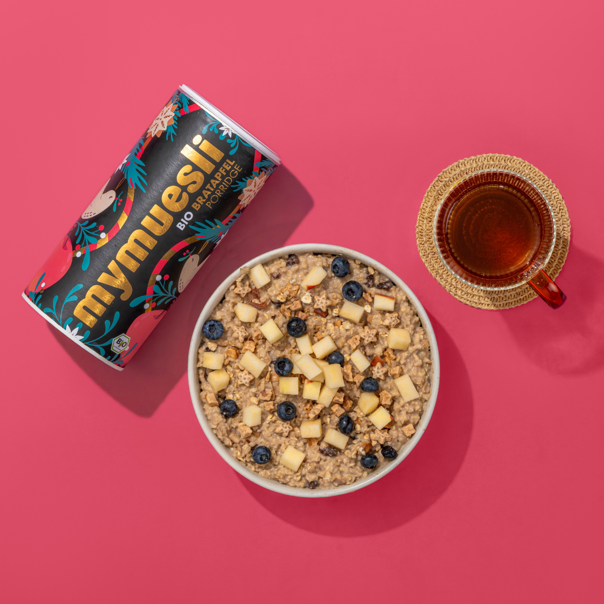 Produktabbildung des mymuesli Bio Bratapfel Porridge. Dazu eine angerichtete Schale mit Bratapfel Porridge, den kleinen Bratapfelsternen, frischen Äpfeln und Blaubeeren. Als Getränk ein Tee. Alles auf einem einfarbigen, hellrotem Hintergrund.