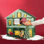 Grünes Adventskalenderhaus mit leichtem Schnee. Vor dem Haus läuft eine Frau mit ihrem Dackel vorbei. Ein Mann steht auf der Leiter und schmückt einen Tannenbaum. Die Fenster des Hauses sind beleuchtet. Eine Frau nimmt einen Müslibecher aus einem geöffneten. 