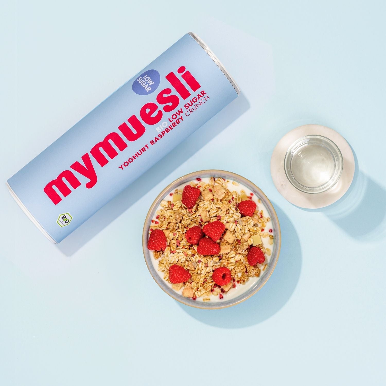 mymuesli Bio Low Sugar Yoghurt Raspberry Crunch Dose mit Müsli in Schale und ein Wasser Glas