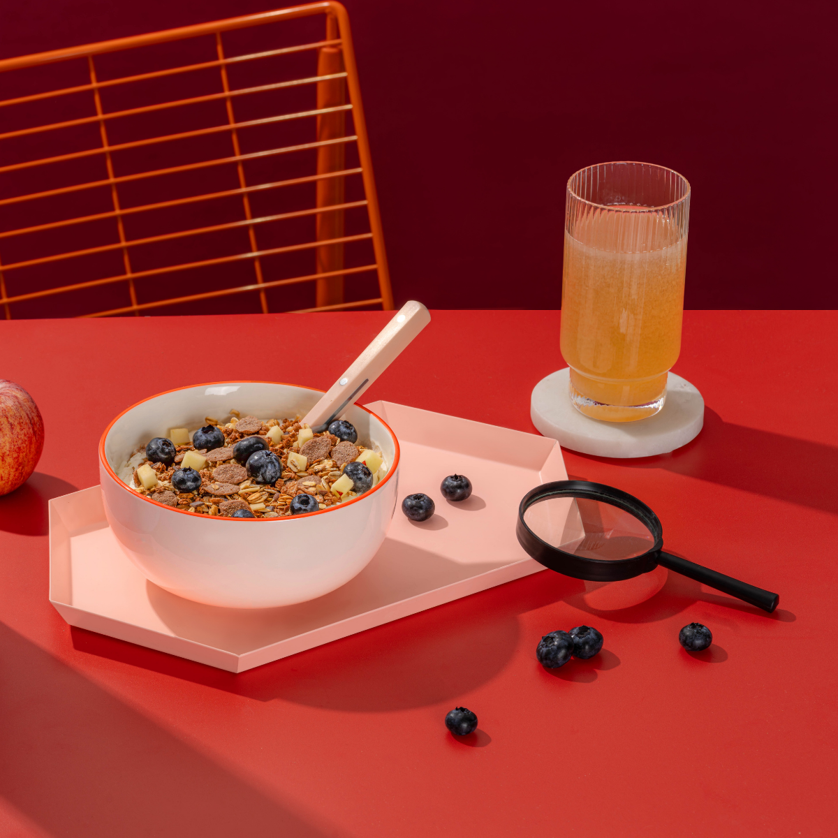 Ein Moodbild des angerichteten mymuesli MicroMacro: Crime City Müslis auf einem Tablett. Garniert mit Apfel und Blaubeeren. Nebenan lieg  eine Lupe für das Suchspiel auf der Verpackung. Dazu eine Apfelschorle im Glas. 