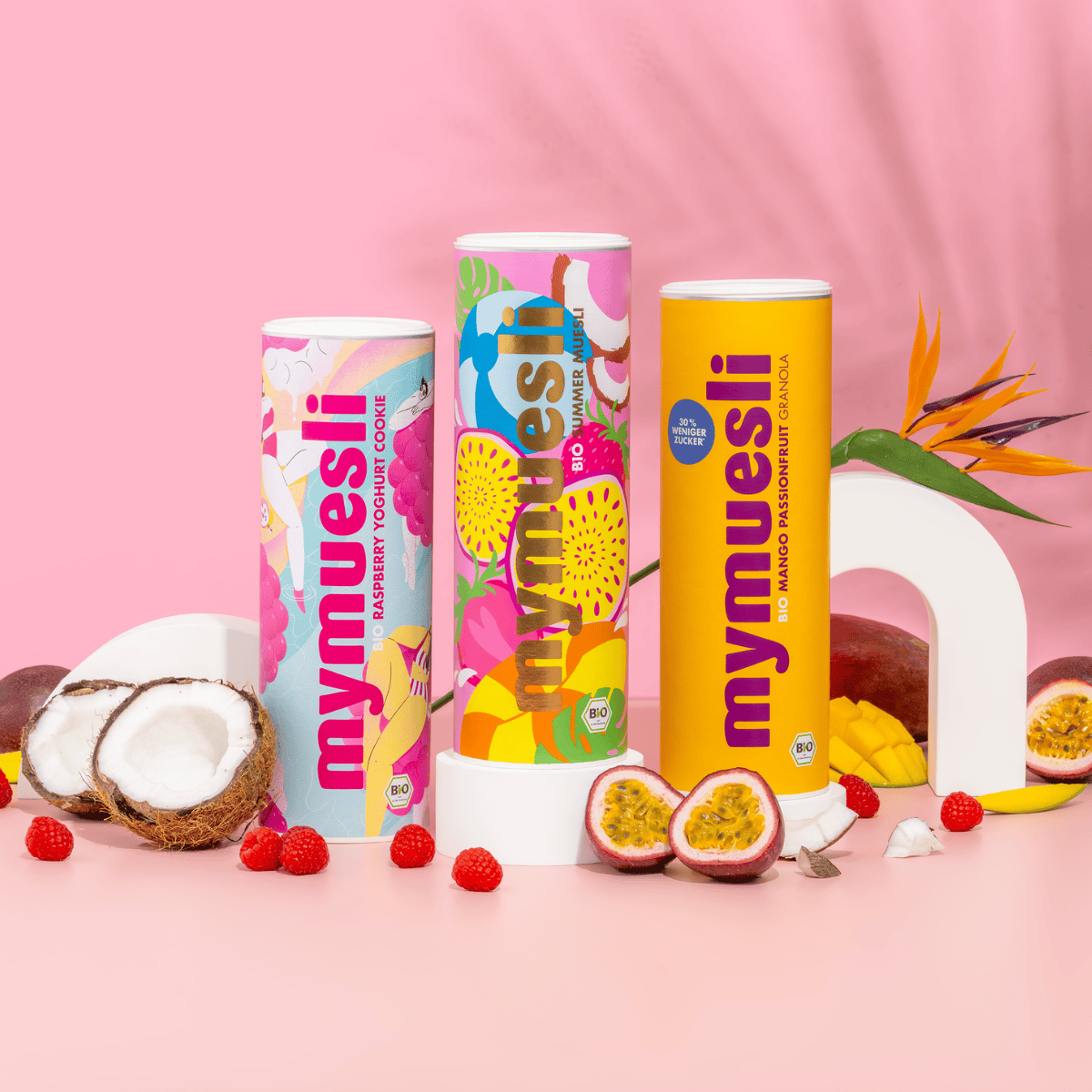 Drei sommerliche Müslidosen sind auf rosa Grund mit Kokosnüssen, Passionsfrüchten und Beeren dekorativ arrangiert.