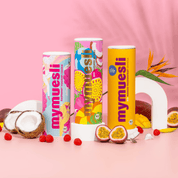 Drei sommerliche Müslidosen sind auf rosa Grund mit Kokosnüssen, Passionsfrüchten und Beeren dekorativ arrangiert.