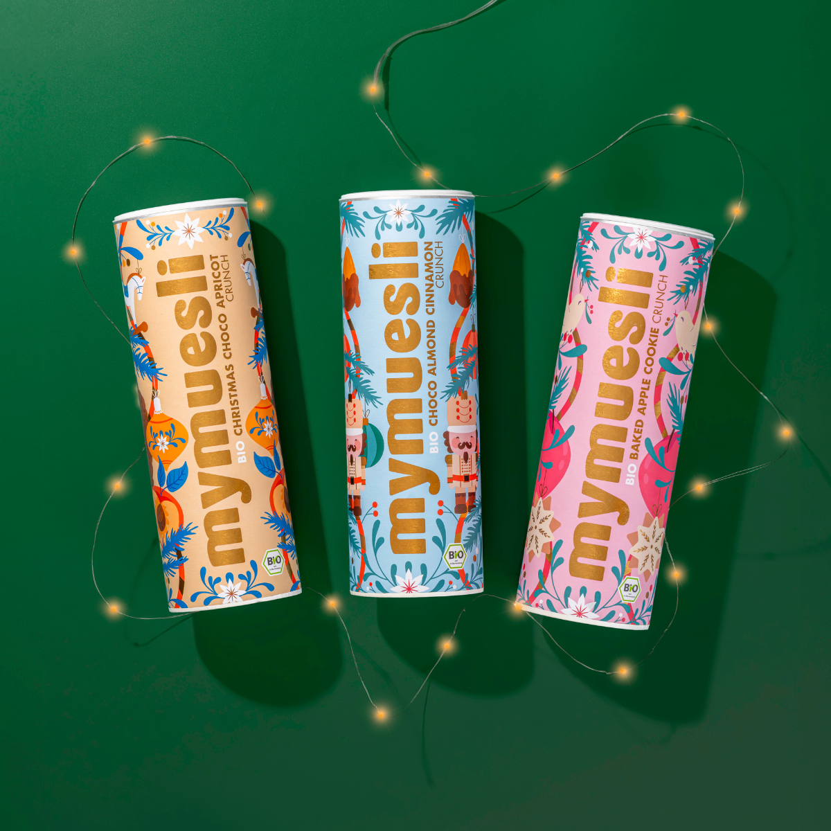 mymuesli Bio Christmas Choco Almond Cinnamon Crunch, Bio Christmas Choco Apricot Crunch und die Sorte Bio Baked Apple Cookie Crunch liegen nebeneinander auf grünen Hintergrund. Verziert sind die Bio Wintermüslis mit einer Lichterkette.