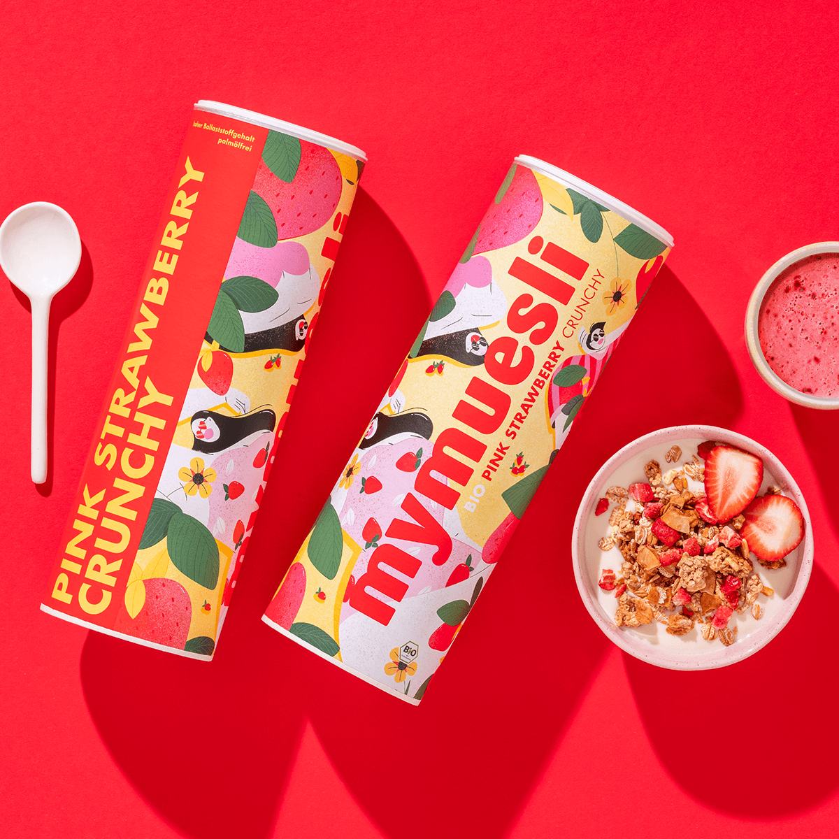 mymuesli Bio Pink Strawberry Crunchy in Schüssel mit Joghurt und frischen Erdbeeren neben Dosen