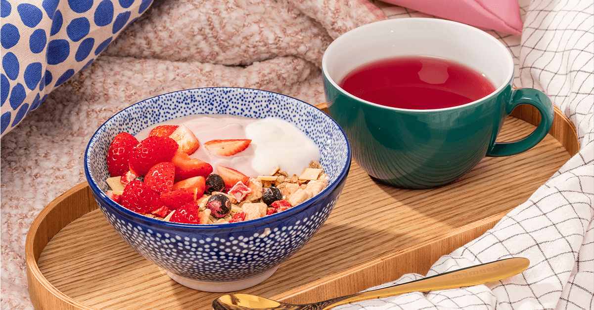 mymuesli Bio Strawberry Cheesecake Müsli in Schüssel mit frischen Erdbeeren und Joghurt auf Tablett mit Tee im Bett