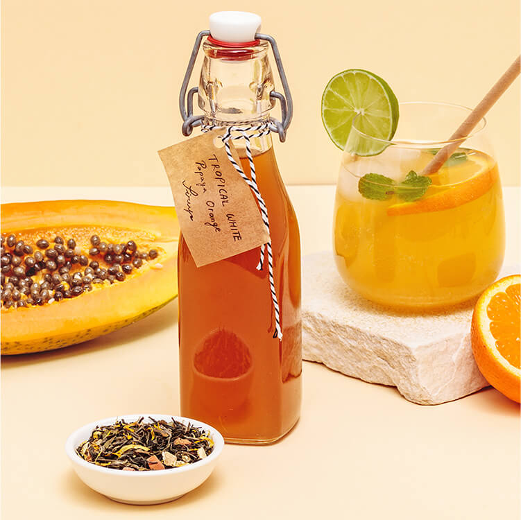 Tropical White Sirup in einer Flasche und in einem Glas, mit Papaya und Orange daneben