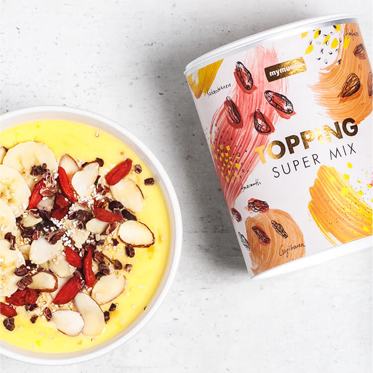 Mango-Kurkuma Bowl in einer Schale mit Bananenscheiben und der Topping Super Mix daneben