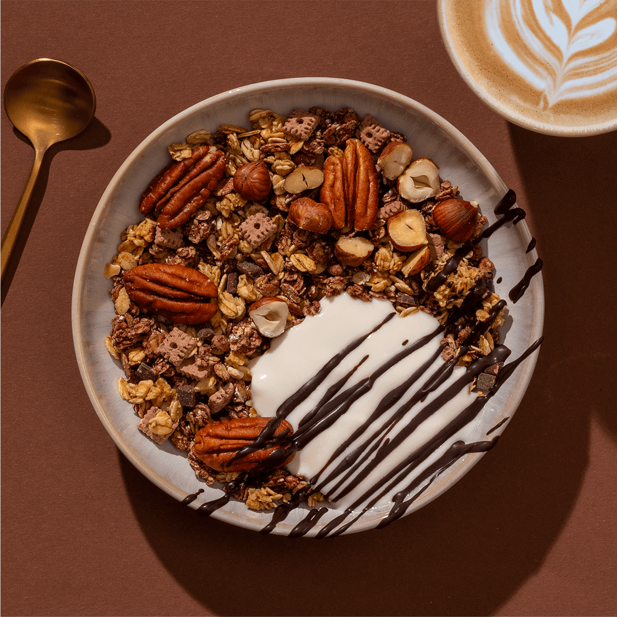 Eine Schüssel Granola mit Joghurt, ganzen Pekannüssen und Schokoladensauce, daneben ein Cappuccino auf braunem Untergrund.