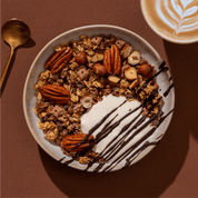 Eine Schüssel Granola mit Joghurt, ganzen Pekannüssen und Schokoladensauce, daneben ein Cappuccino auf braunem Untergrund.