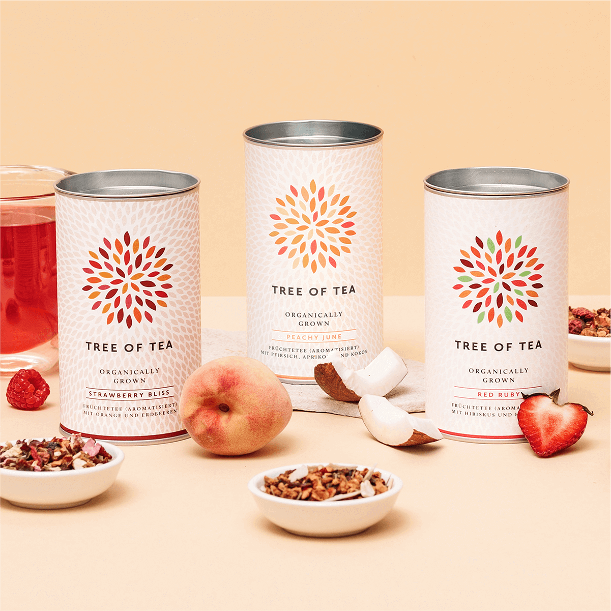 Tree of Tea Bio Früchteteeset mit Red Ruby, Peachy June und Strawberry Bliss Tee neben Teeglas, Pfirsich, Kokosnuss und Erdbeeren