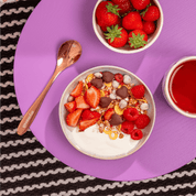 Frühstücks-Ensemble auf lila Untergrund: Eine Joghurt-Bowl mit Erdbeeren und Schokomüsli, dazu ein kupferfarbener Löffel und frische Erdbeeren.