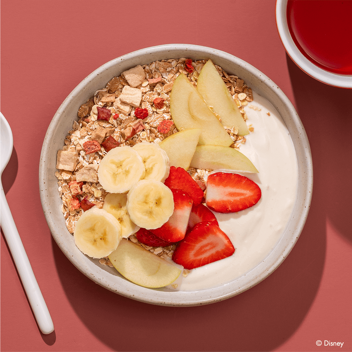 Eine helle Schüssel mit weißem Joghurt oder Quark, getoppt mit Müsli, Bananenscheiben, Erdbeerstücken und Apfelspalten auf rötlichem Untergrund.