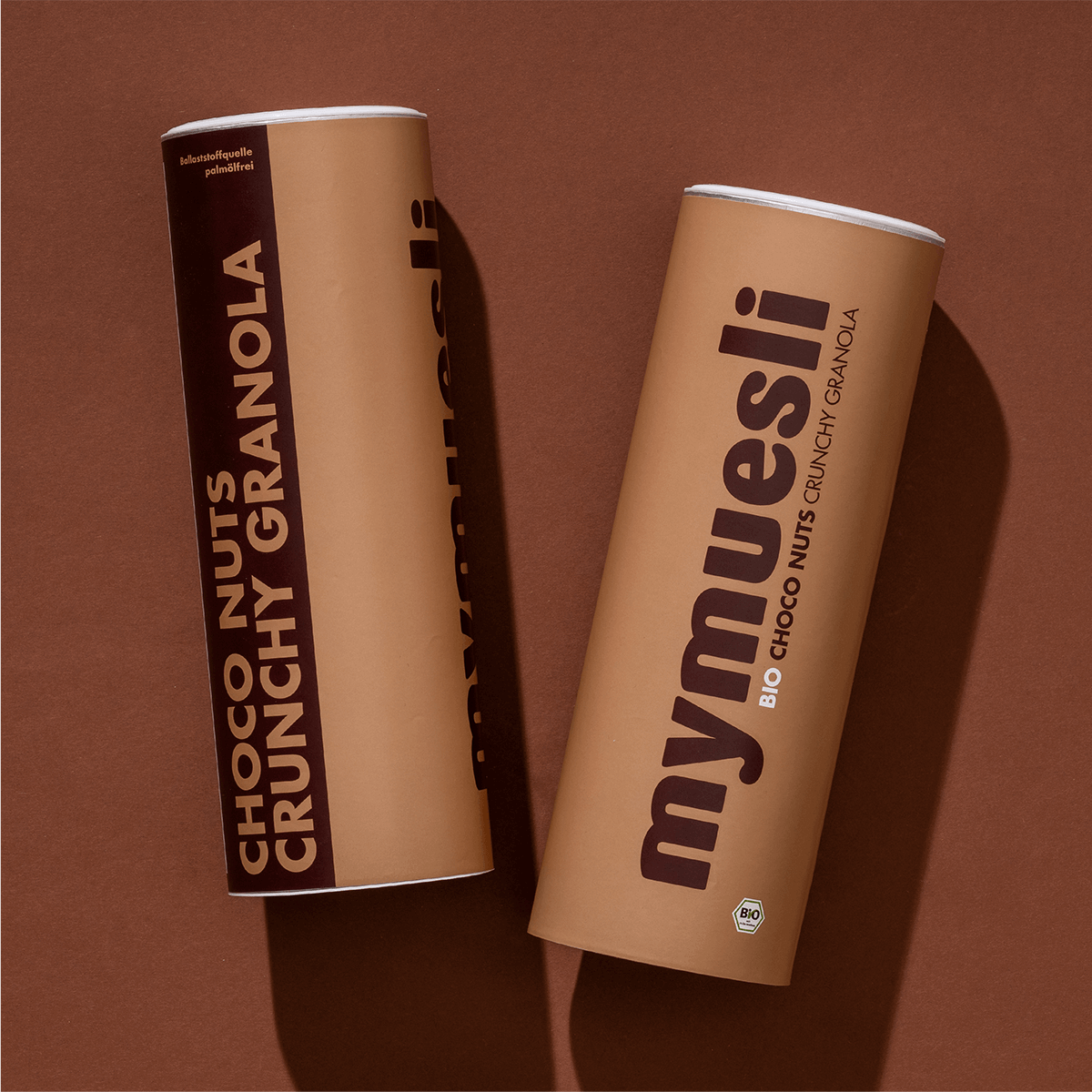 Zwei beigefarbene mymuesli -Dosen der Sorte "Choco Nuts Crunchy Granola" liegen nebeneinander auf einem braunen Hintergrund.