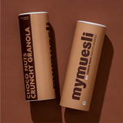 Zwei beigefarbene mymuesli -Dosen der Sorte "Choco Nuts Crunchy Granola" liegen nebeneinander auf einem braunen Hintergrund.