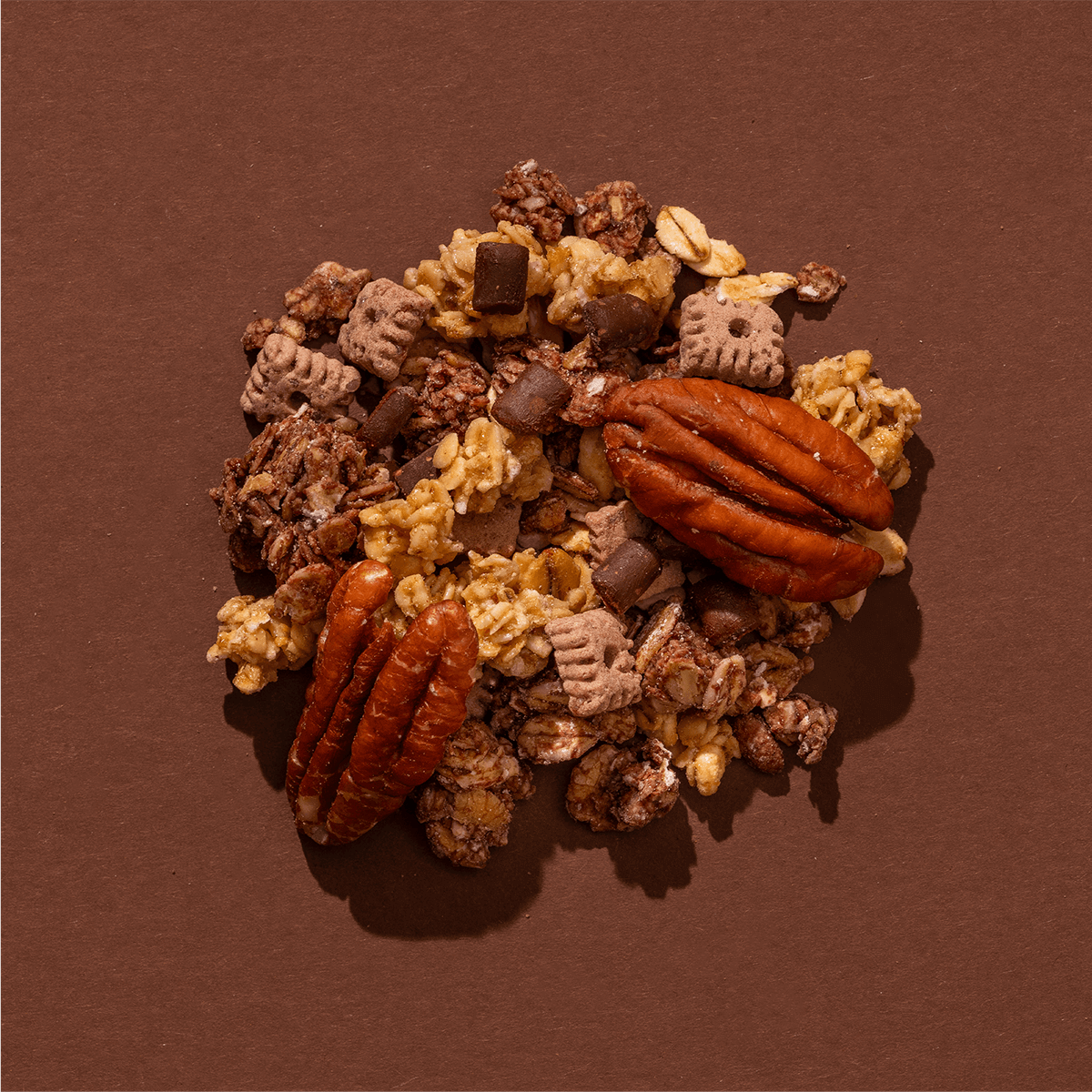 Eine Nahaufnahme der Granola-Mischung auf braunem Grund mit großen Pekannüssen, Knusper-Clustern und Schokoladenstücken.