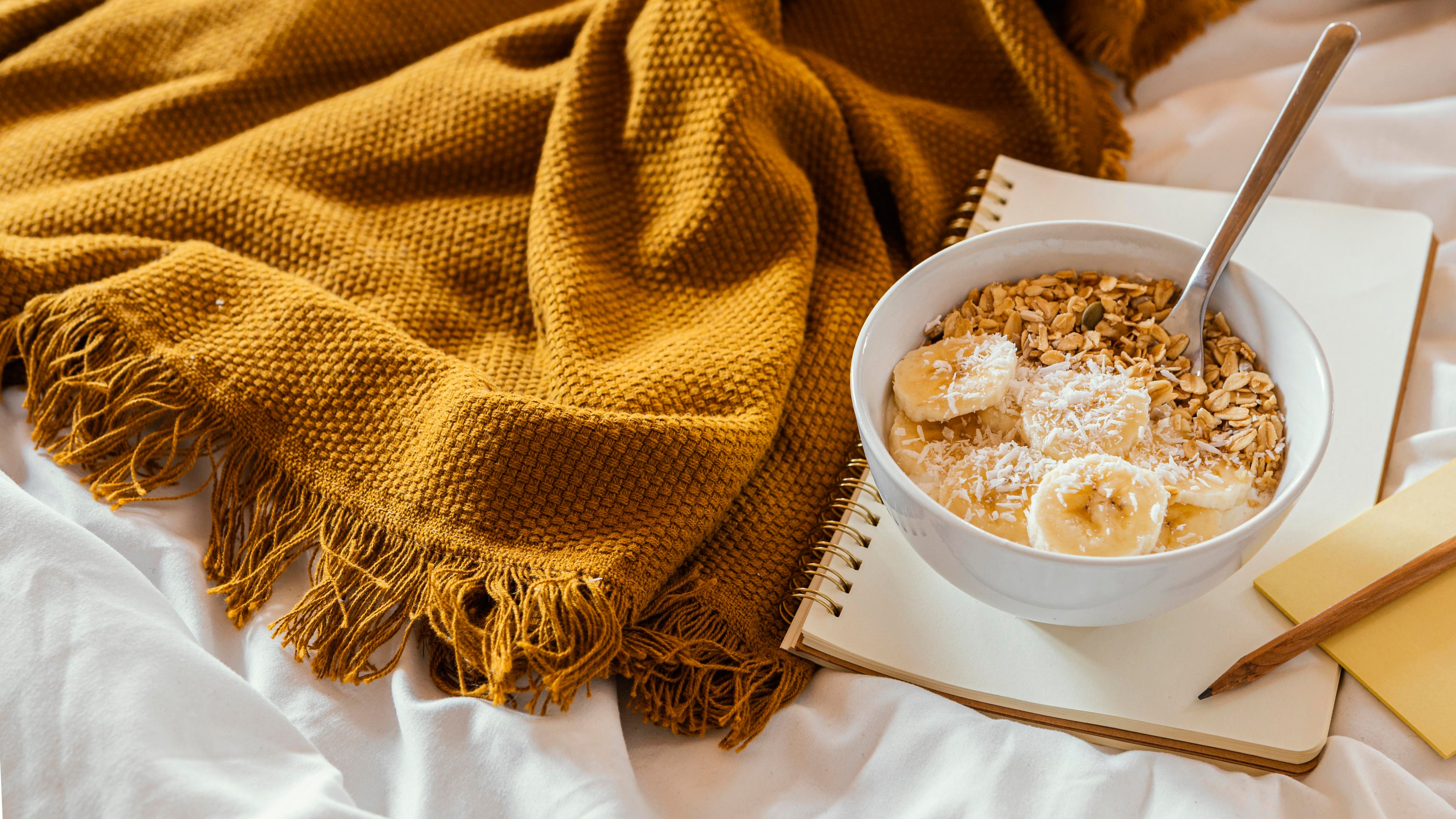 Eine angerichtete Müsli Schale mit Müsli, frischen Bananen und Kokosraspeln. Die SChale steht auf einem Notizblock, daneben eine weiche, geld-braun-farbende Decke. 