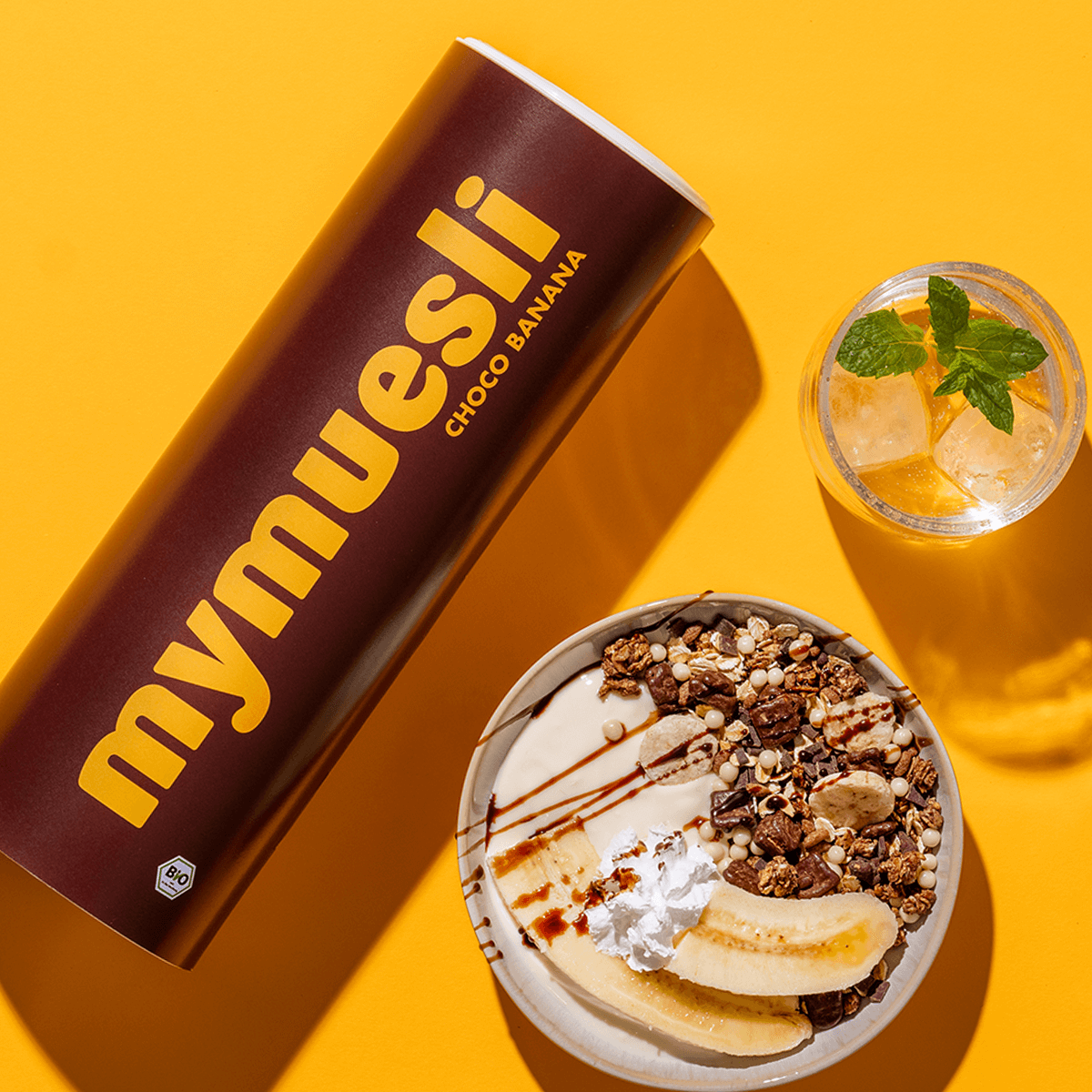 mymuesli Bio Choco Banana Müsli Dose und daneben eine Schale mit Choco Banana Müsli, Joghurt, zwei halbe Bananen und Schokosauce