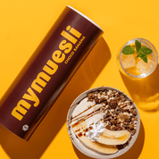 mymuesli Bio Choco Banana Müsli Dose und daneben eine Schale mit Choco Banana Müsli, Joghurt, zwei halbe Bananen und Schokosauce