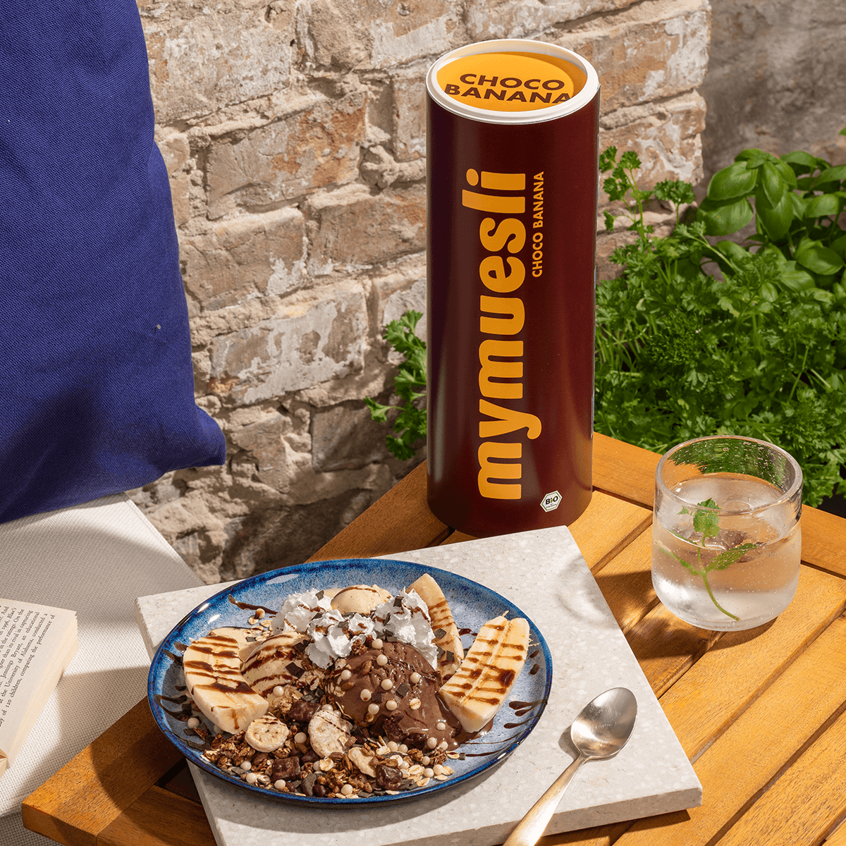 mymuesli Bio Choco Banana Müsli Dose und daneben eine Schale mit Choco Banana Müsli, Joghurt, zwei halbe Bananen und Schokosauce