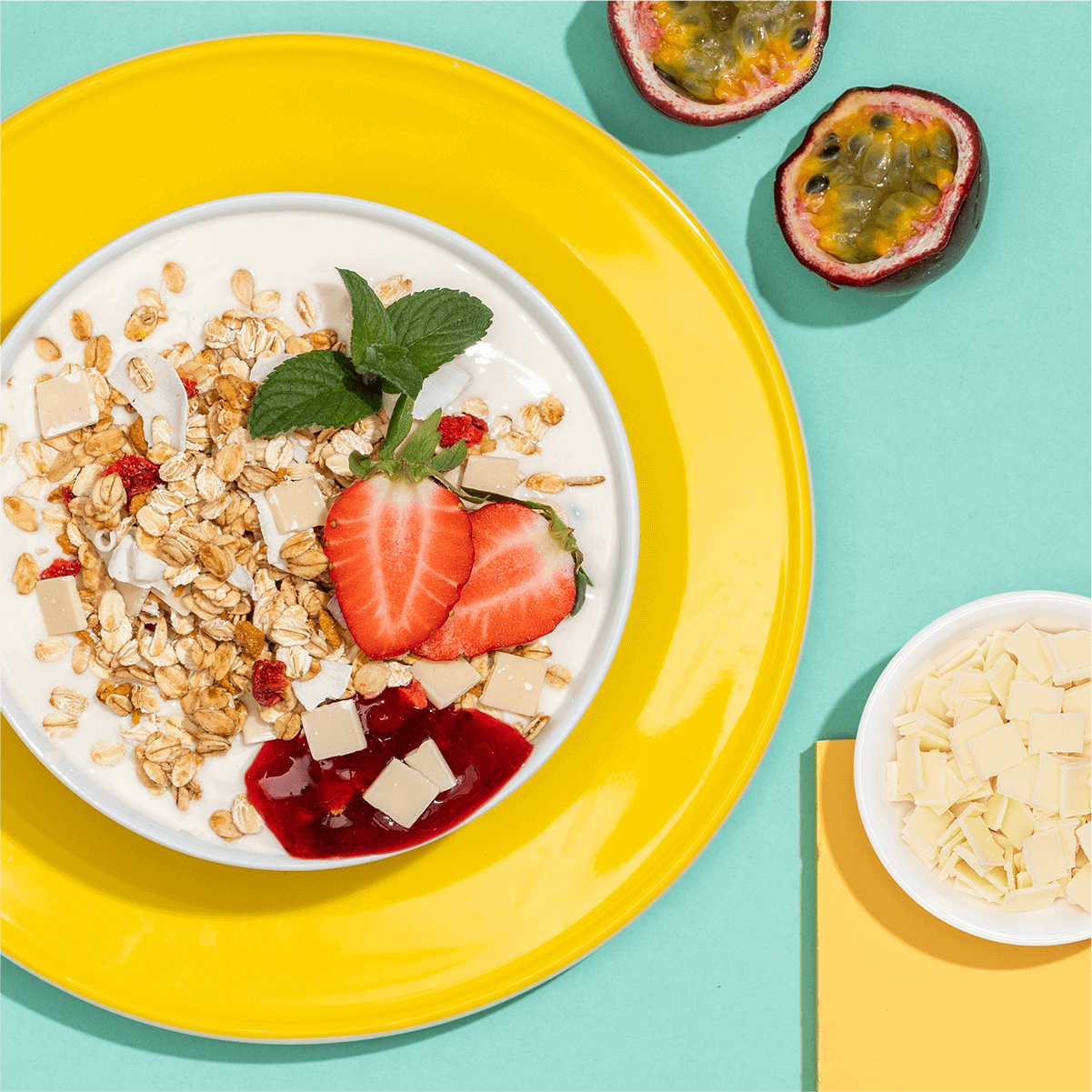 Volle Mueslischale, Maracujas und Joghurtplaettchen auf einem gelb und tuerkis gestalteten Tisch