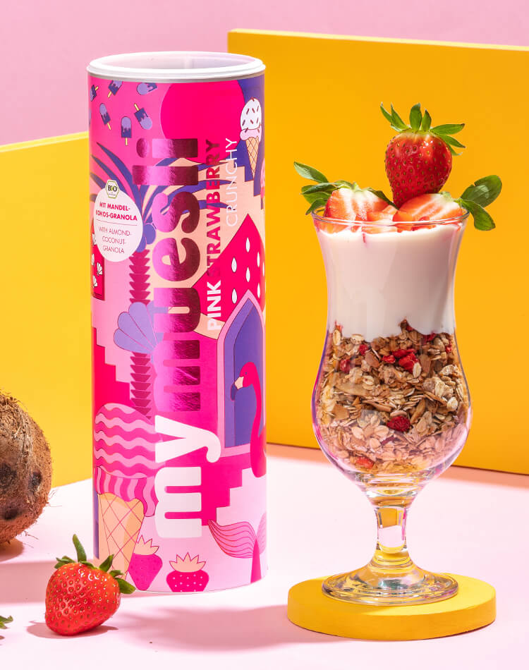 Pink Strawberry Crunchy Muesli Dose mit Flamingo Motiv neben einem Cocktailglas gefuellt mit Muesli, Joghurt und Erdbeeren