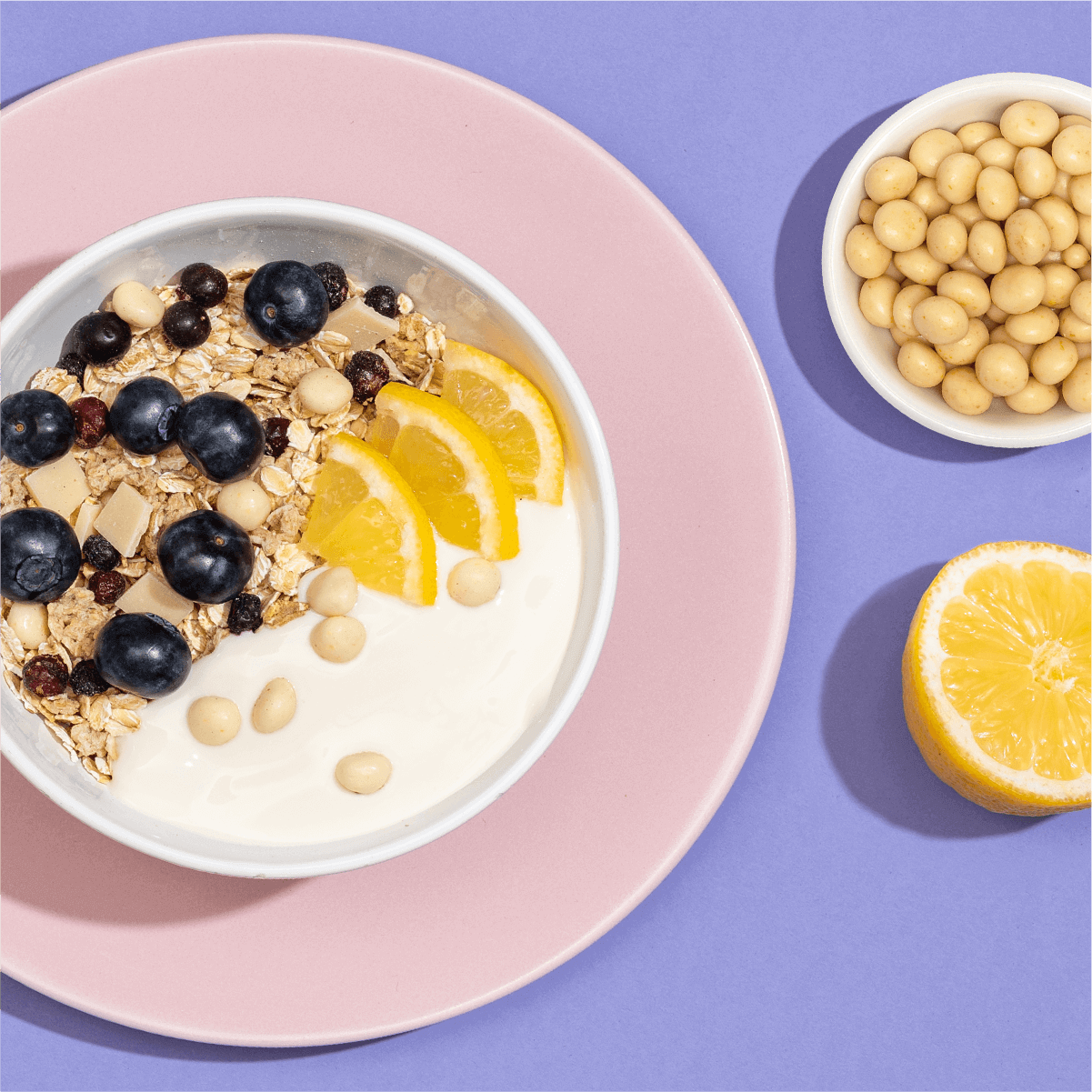 Mueslischale mit Joghurt, Zitrone und Heidelbeeren vor einem hellen lila Hintergrund