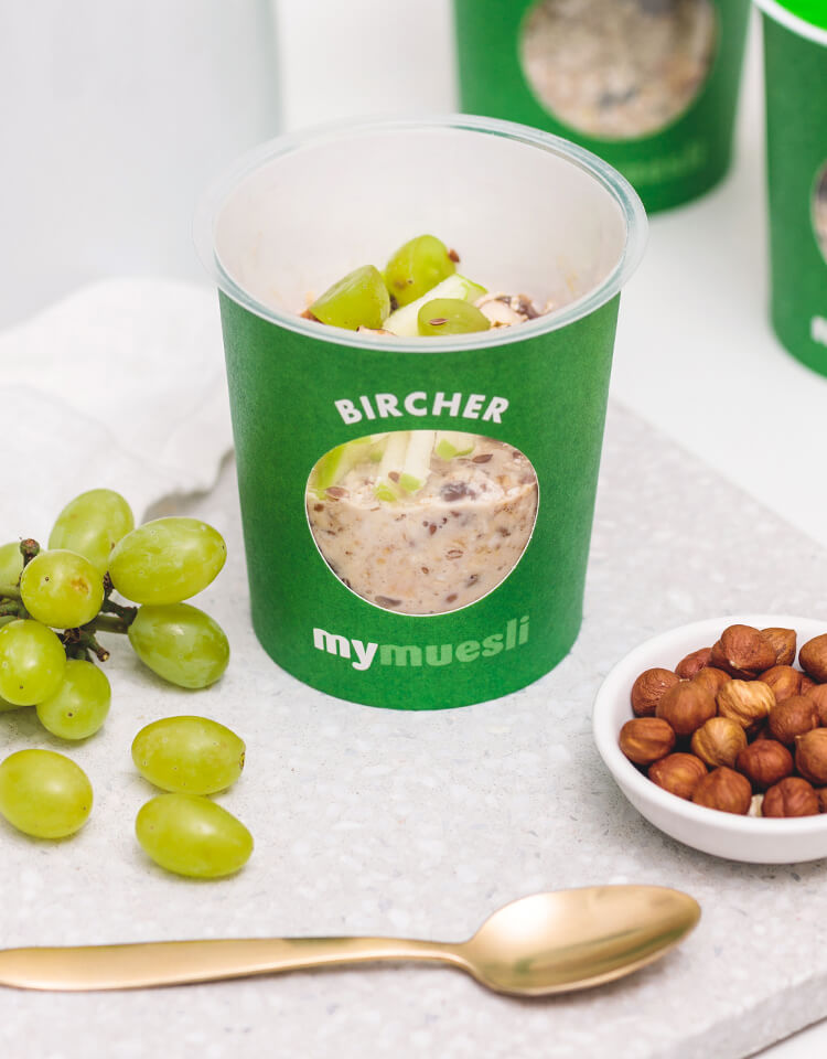 Gruener mymuesli Bircher To-Go-Becher, daneben ein goldener Muesliloeffel, Weintrauben und Haselnuesse