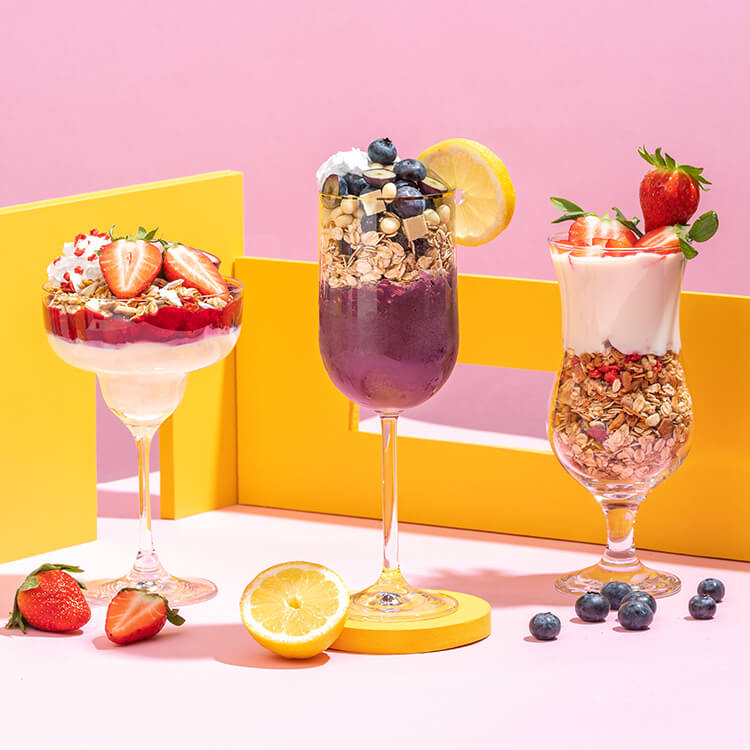 Drei Cocktailgläser mit Muesli Desserts gefuellt vor sommerlich hellem Hintergrund