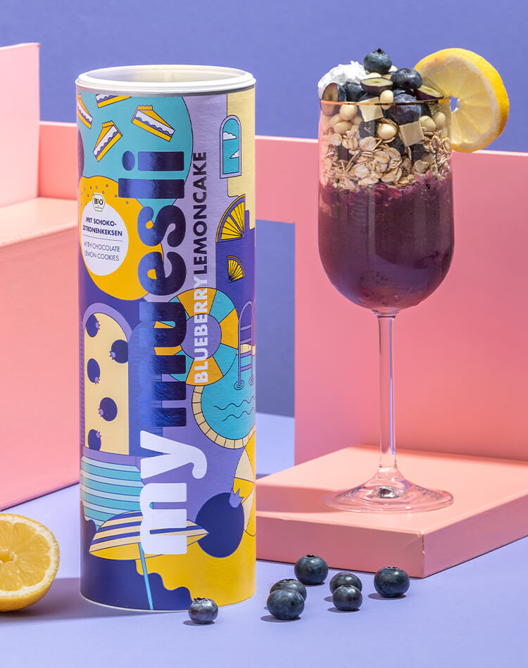 Mueslidose mit der Sorte Blaubeeren Zitronenkuchen neben Cocktailglas mit Mueslidessert