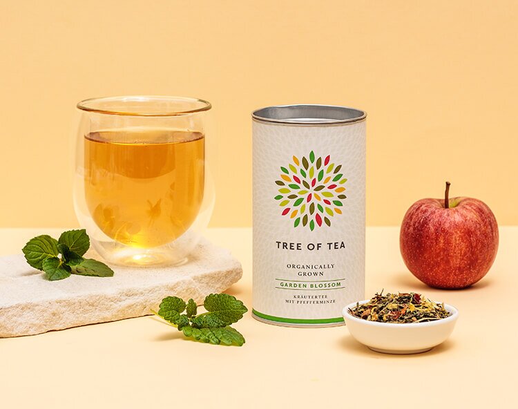 Teedose von Tree of Tea mit Teeglas, frischer gruener Minze und einem roten Apfel